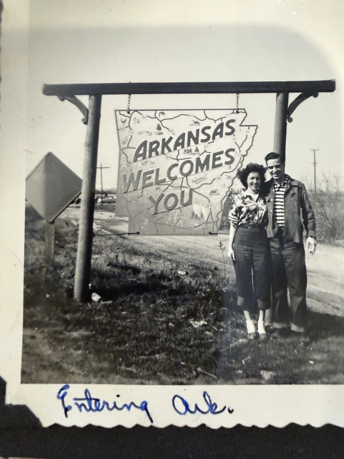 Two people standing beside an Arkansas Welcomes You sign, with the handwritten caption “Entering Ark.”