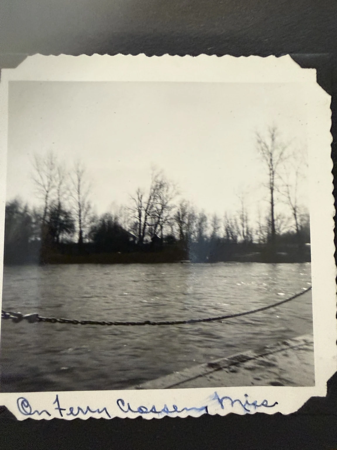 The river and opposite bank viewed across a ferry’s safety chain, with a handwritten Mississippi-crossing caption.