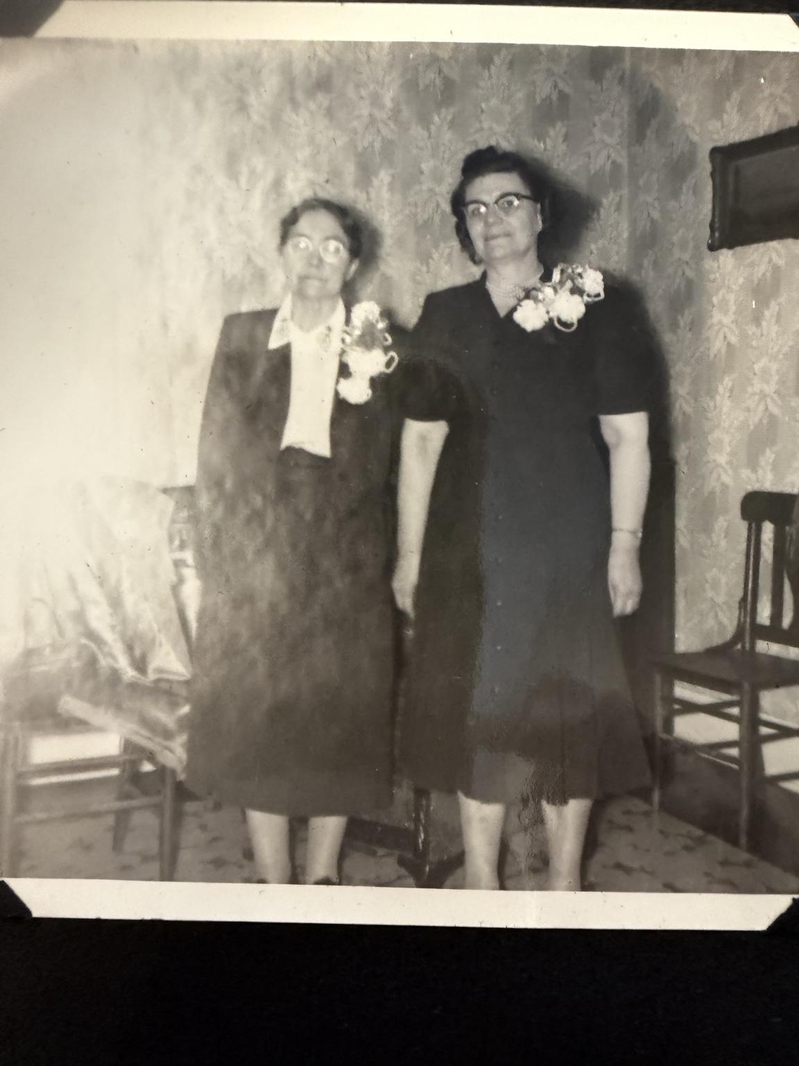 Two women with corsages posed beside chairs against patterned wallpaper.
